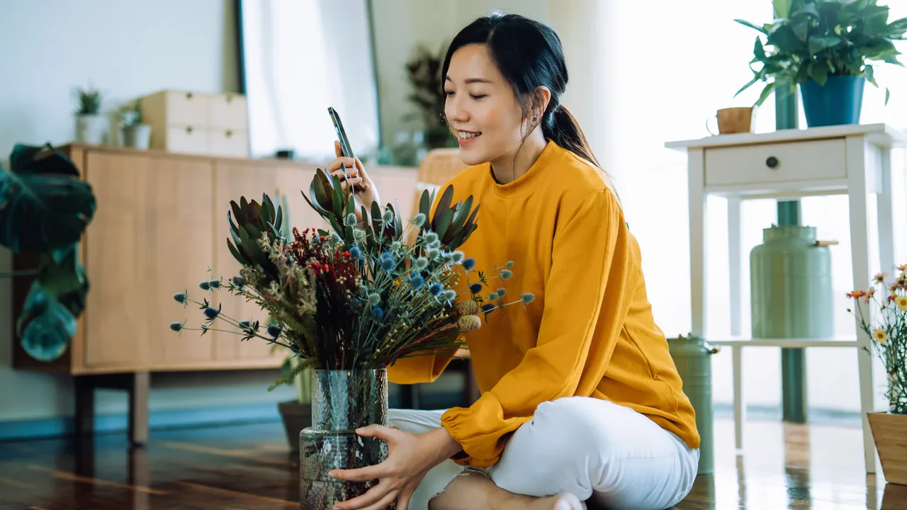 A woman holding a smartphone beside a vase of flowers in a bright room.