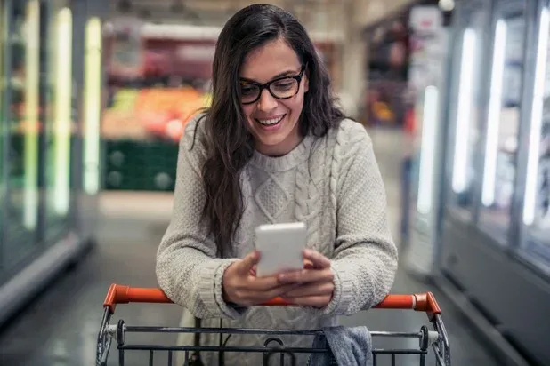 A shopper holding a smartphone behind a grocery cart in the illustrative image supplied for BHN's Drive and Grocery card announcement.