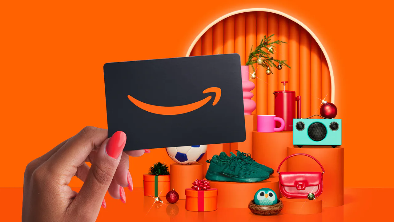 A hand holds a dark Amazon gift card in front of an orange display of gifts and holiday decorations.