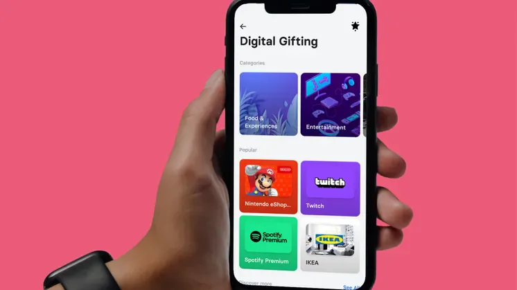 A hand holds a phone displaying Revolut's Digital Gifting screen with gift card brands on a pink background.