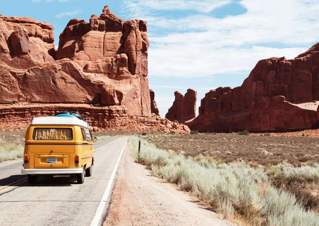A yellow camper van travels along a road between red sandstone cliffs beneath a blue sky.