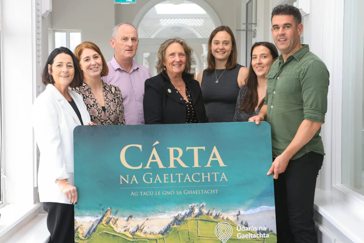 Seven people stand behind a large Cárta na Gaeltachta display card in an official programme photograph.