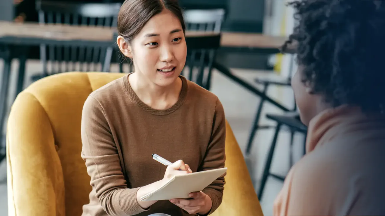 A woman holding a notebook and pen speaks with another person in a seating area.