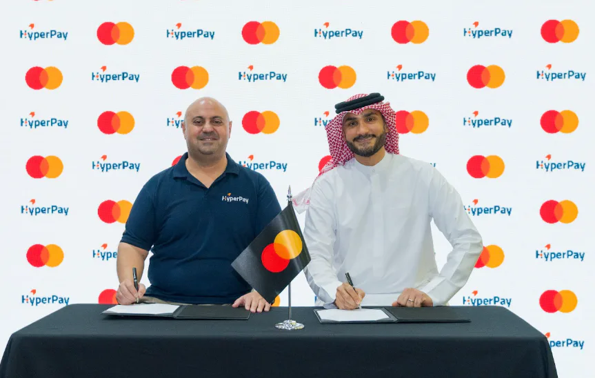 Two men sign documents at a table in front of a backdrop displaying HyperPay and Mastercard logos.