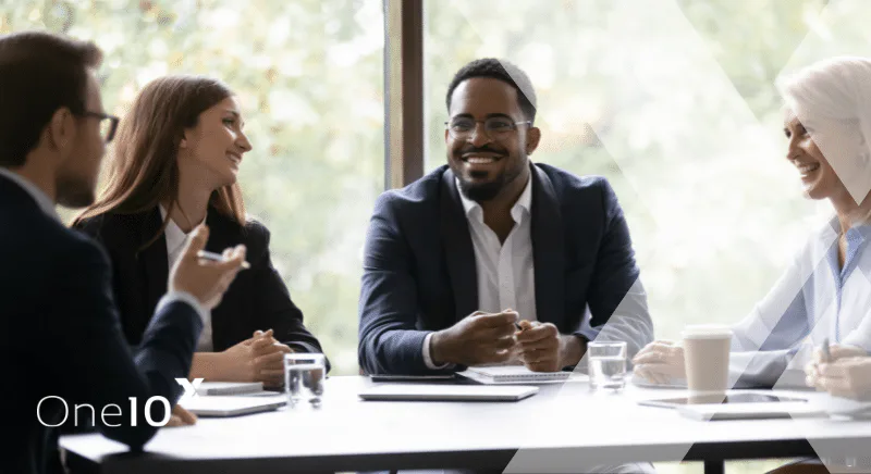 Four Diverse Professionals Share a Friendly Business Meeting Around a Table, with Notebooks and Drinks, One10 Logo Visible in the Corner.