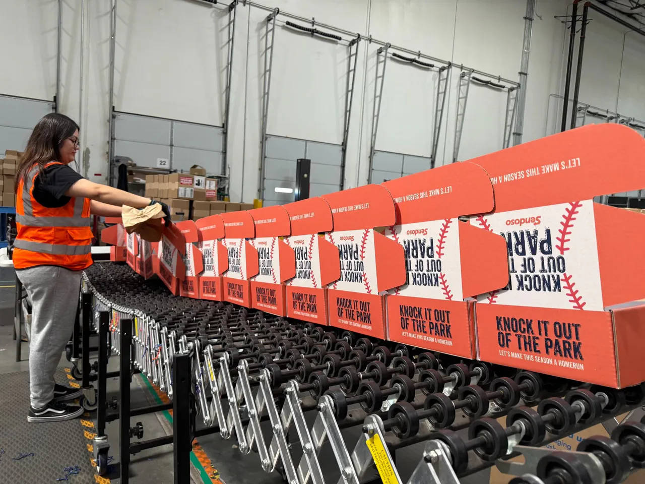 A worker assembles red baseball-themed gift boxes on a warehouse conveyor