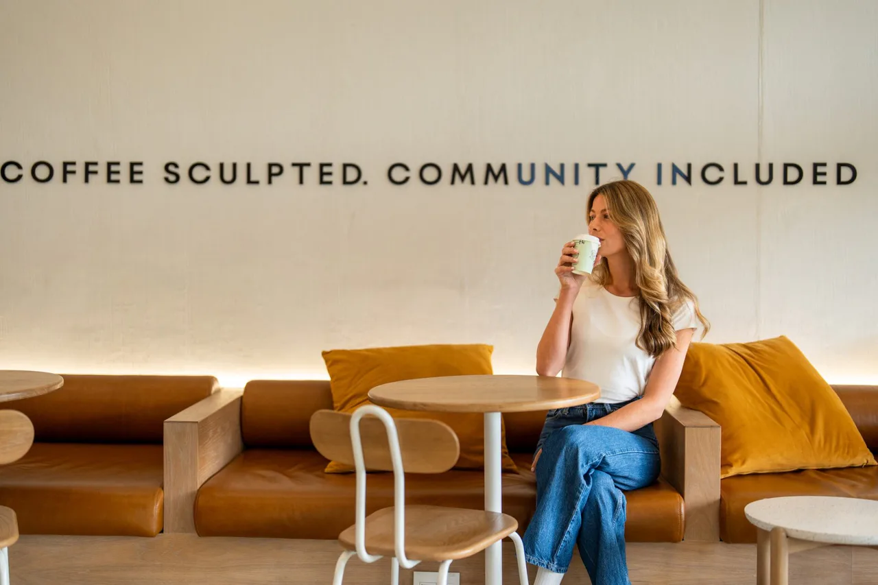 A woman drinking from a takeaway coffee cup while seated at a table inside a Platō café.